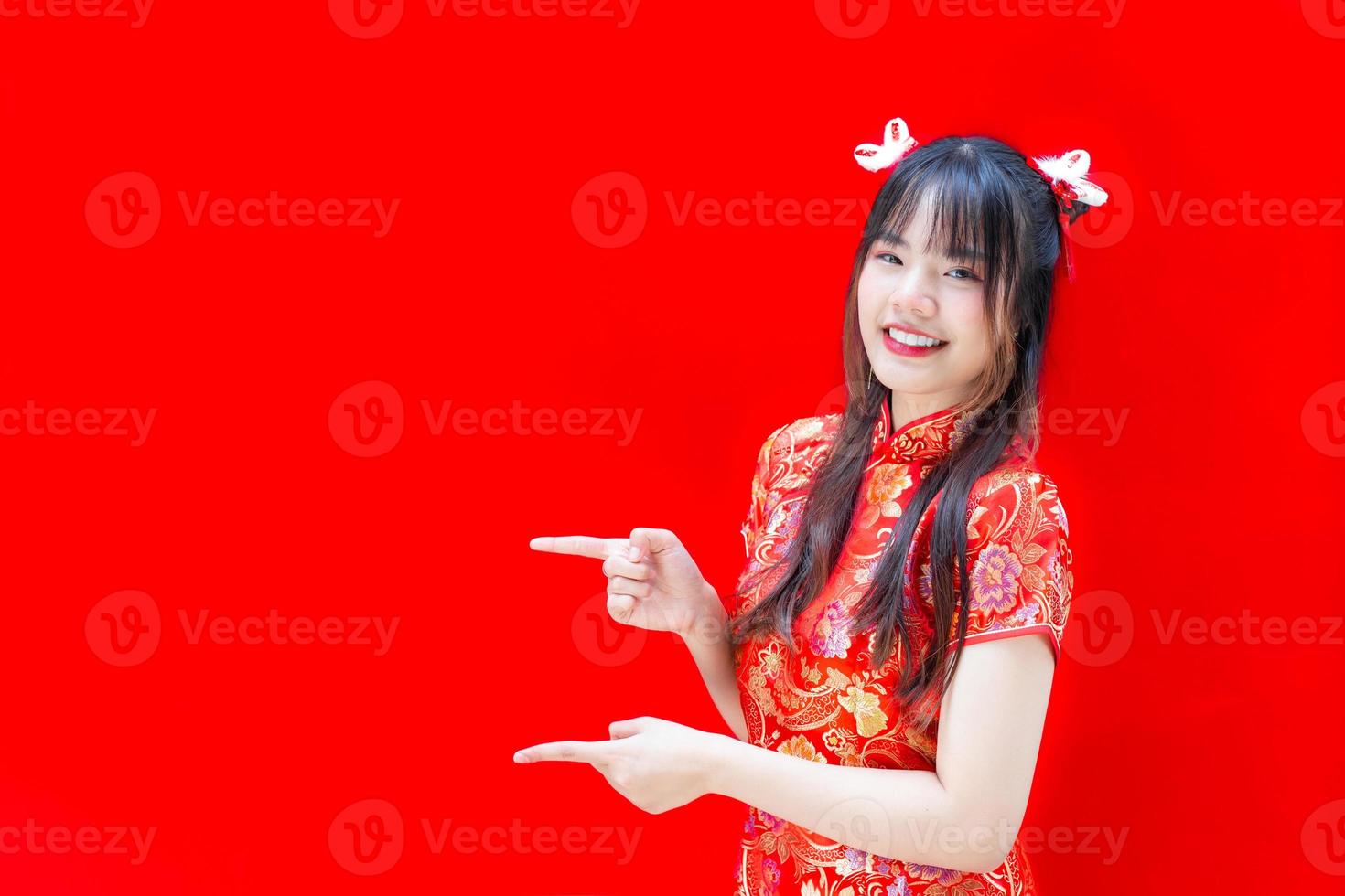 linda chica asiática con el pelo largo que usa un vestido rojo cheongsam en el tema del año nuevo chino mientras muestra su mano para presentar algo sobre un fondo rojo. foto