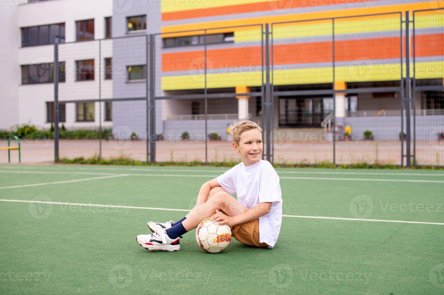 A teenage boy is sitting on a green field in the school yard with a soccer ball photo