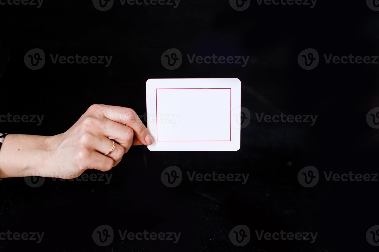 The hand with the ring holds a white card with red lines against a black wall photo