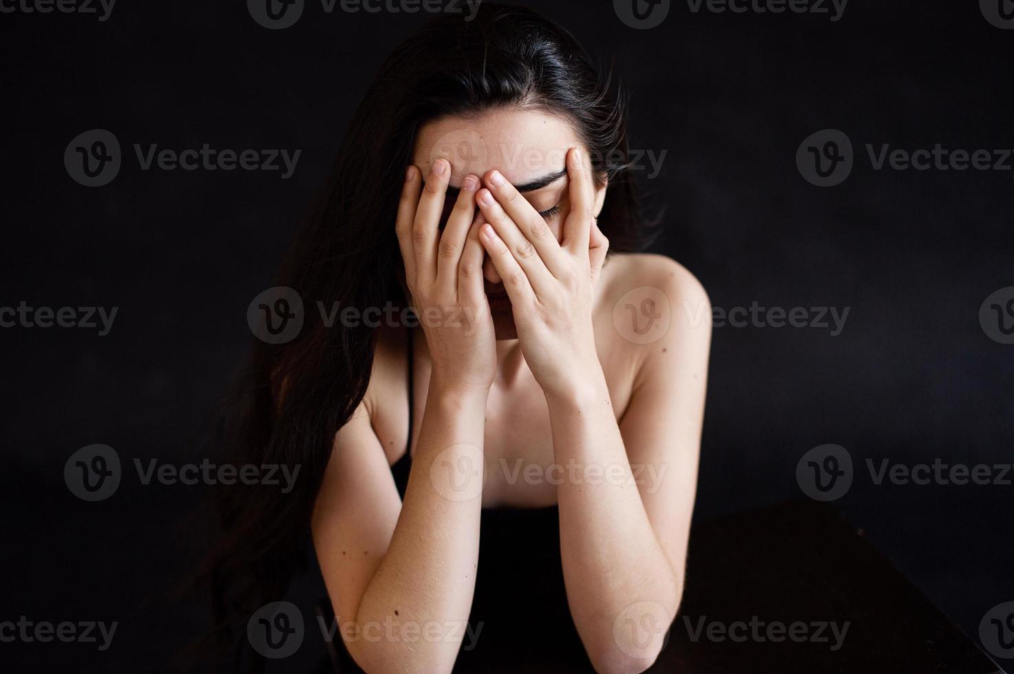 The girl is sitting on a black background, covering her face with her hands photo