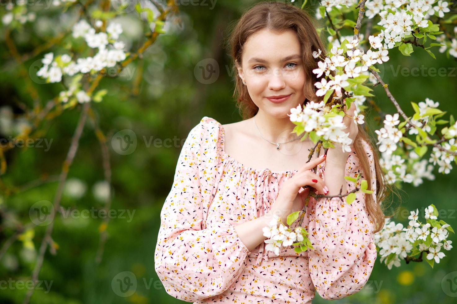 A girl in a pink dress stands near a white tree with flowers looks at the camera and smiles photo