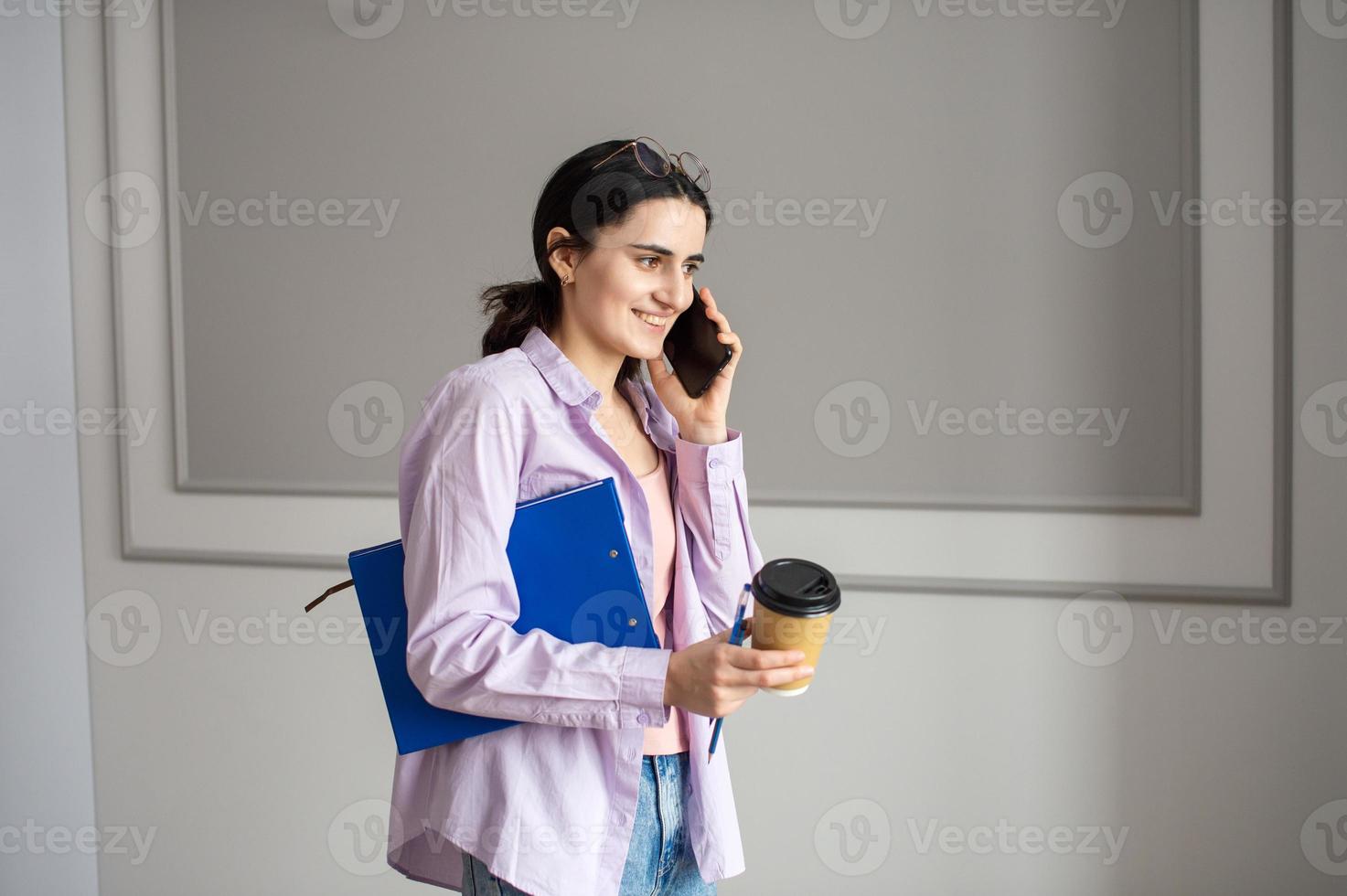 A cheerful girl is talking on the phone and holding a folder with documents and a cup photo