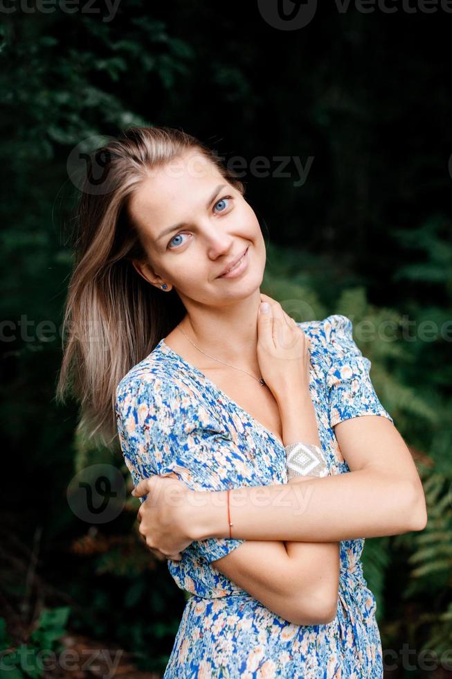 A girl in a blue dress stands in a green forest and holds her neck with her hands photo