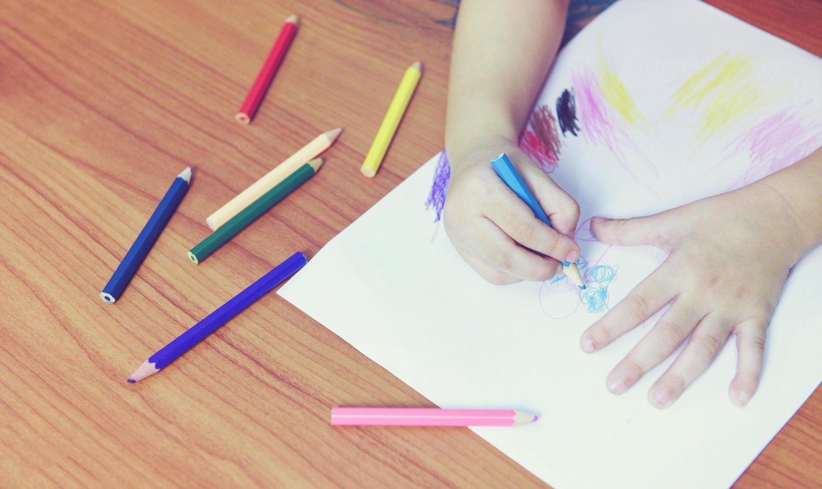 Girl painting on paper sheet with colour pencils on the wooden table at home - child kid doing drawing picture and colorful crayon photo