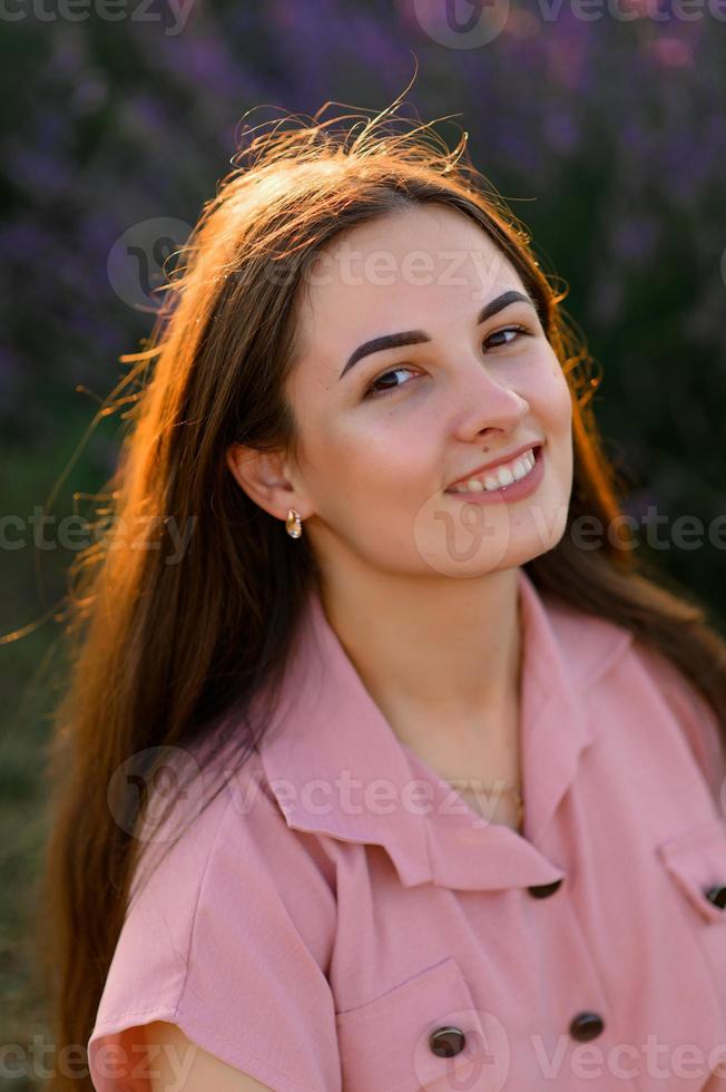 A cheerful young girl in a pink dress and a straw hat in her hands stands among lavender bushes. Sunset. photo