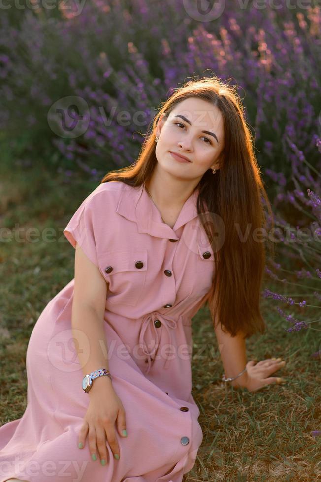 un alegre joven niña en un rosado vestir y un Paja sombrero en su manos soportes entre lavanda arbustos puesta de sol. foto