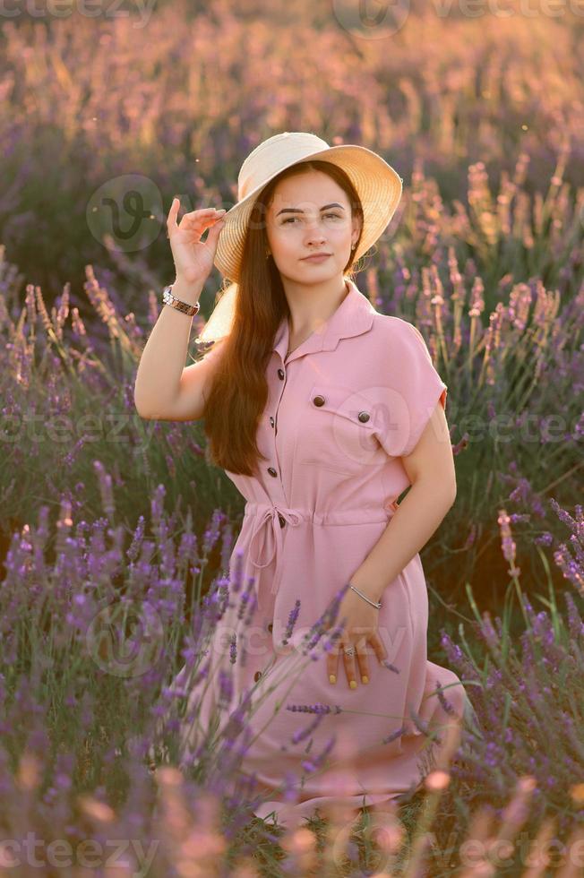 A cheerful young girl in a pink dress and a straw hat in her hands stands among lavender bushes. Sunset. photo