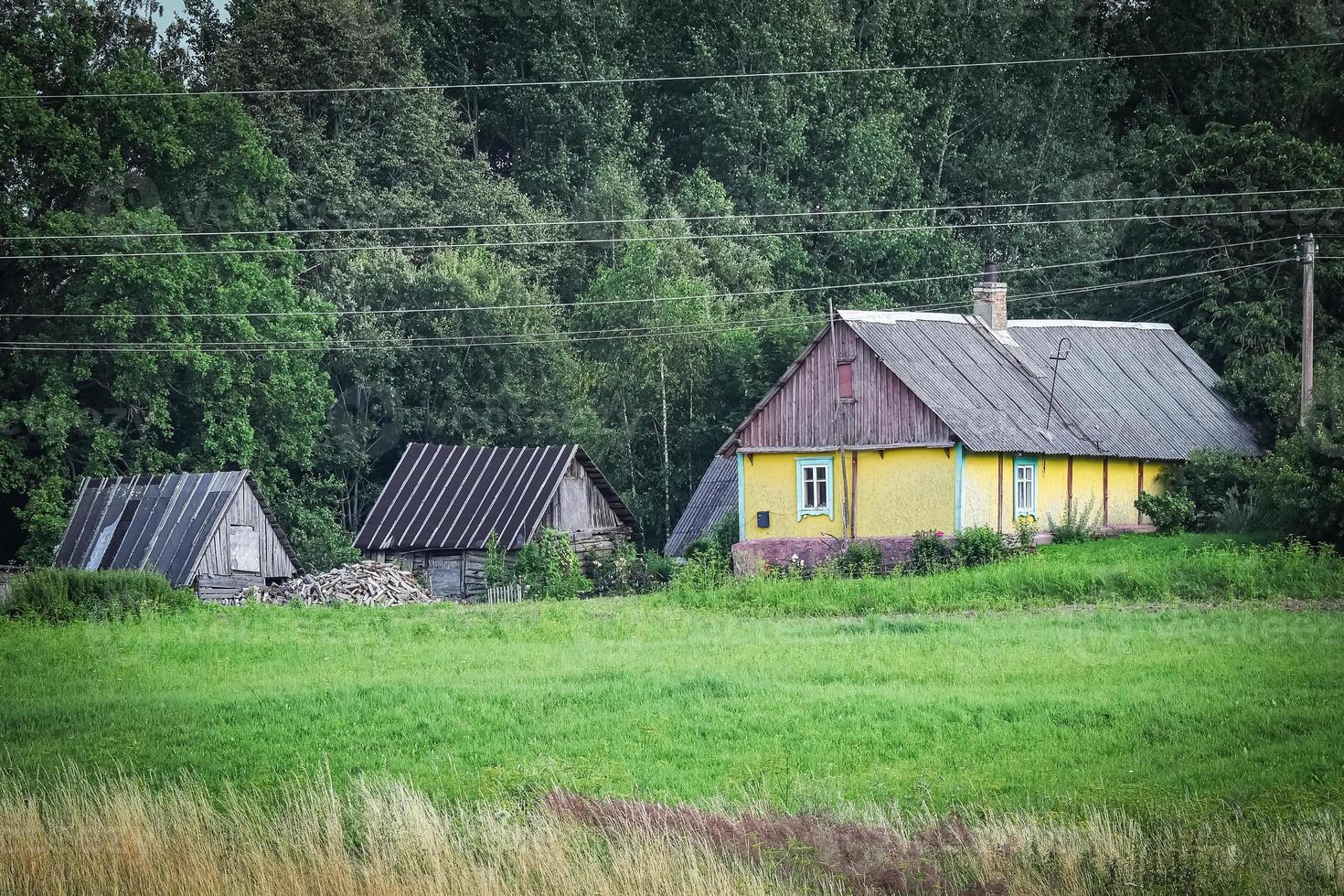 Small yellow farm house in typical surroundings of forest and grass meadow with visible field before harvest in Europe photo