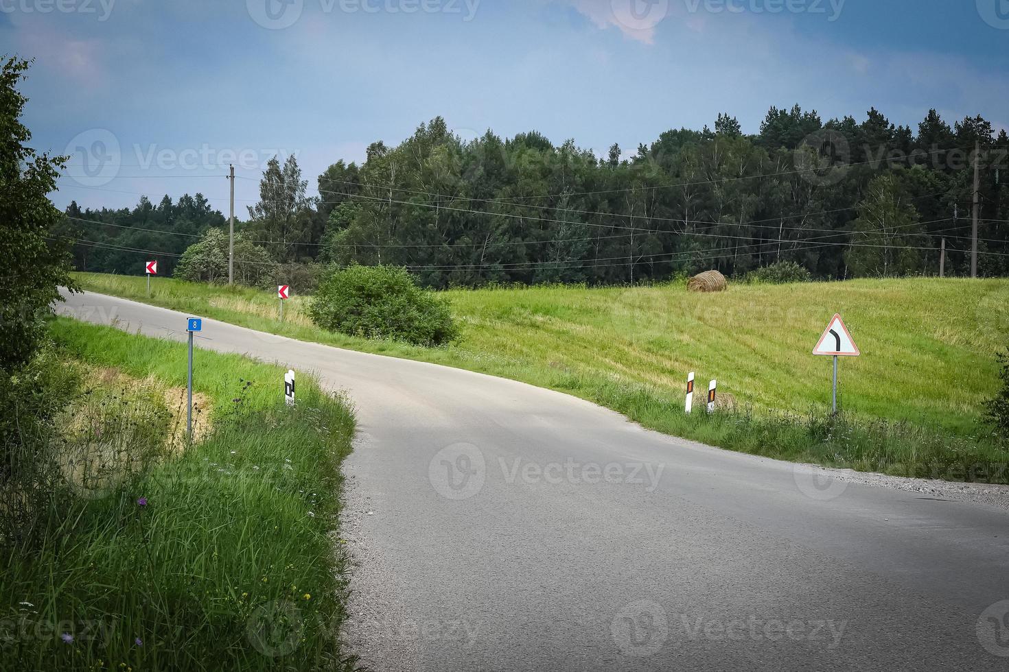 Summer asphalt road without lines but with road signs in green meadows surrounded by forest on sunny day photo