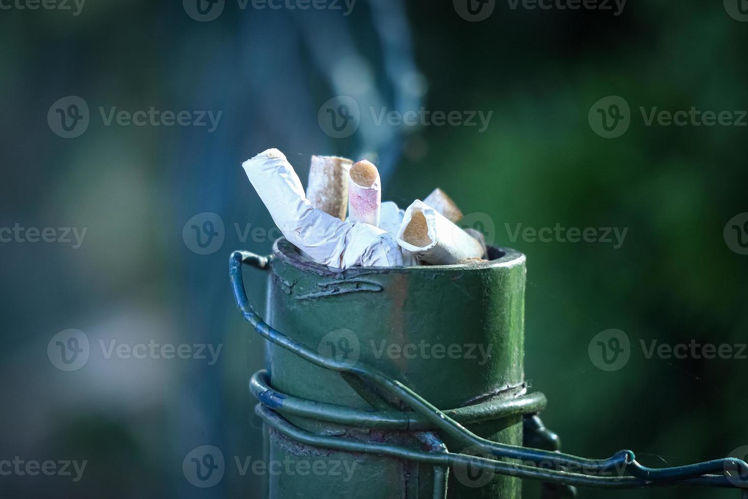 Ashtray made from the street dark green fence pole with visible fence wire with cigarette stubs inside on green blurred background photo