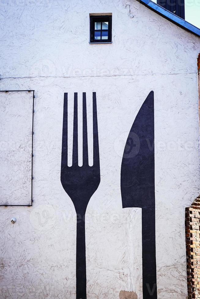 Huge fork and a knife painted in black on white building wall backside with window photo