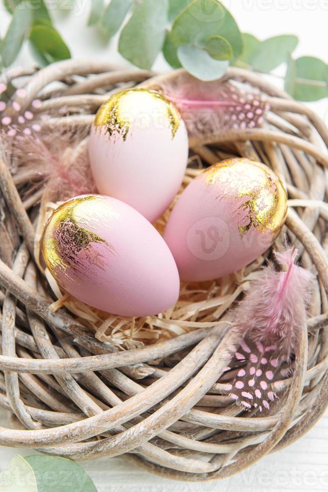 Easter eggs painted pink and gold and  feathers in a nest on a white wooden background. photo