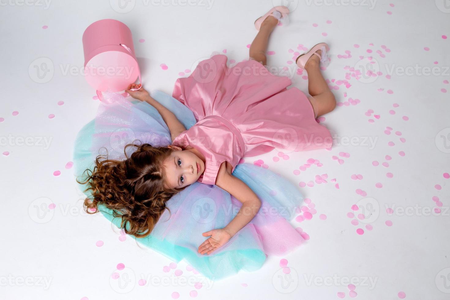 hermosa pequeño niña en un elegante rosado vestir mentiras en el piso esparcido con papel picado. parte superior vista. Moda y estilo. niño celebra su cumpleaños. foto en el estudio. espacio para texto. alto calidad foto