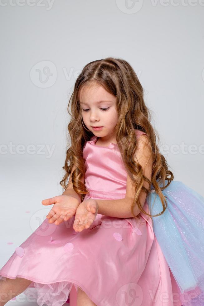 beautiful little girl in a chic pink dress poses sitting on a gray background and throws confetti. fashion and style. space for text. photo in the studio. High quality photo