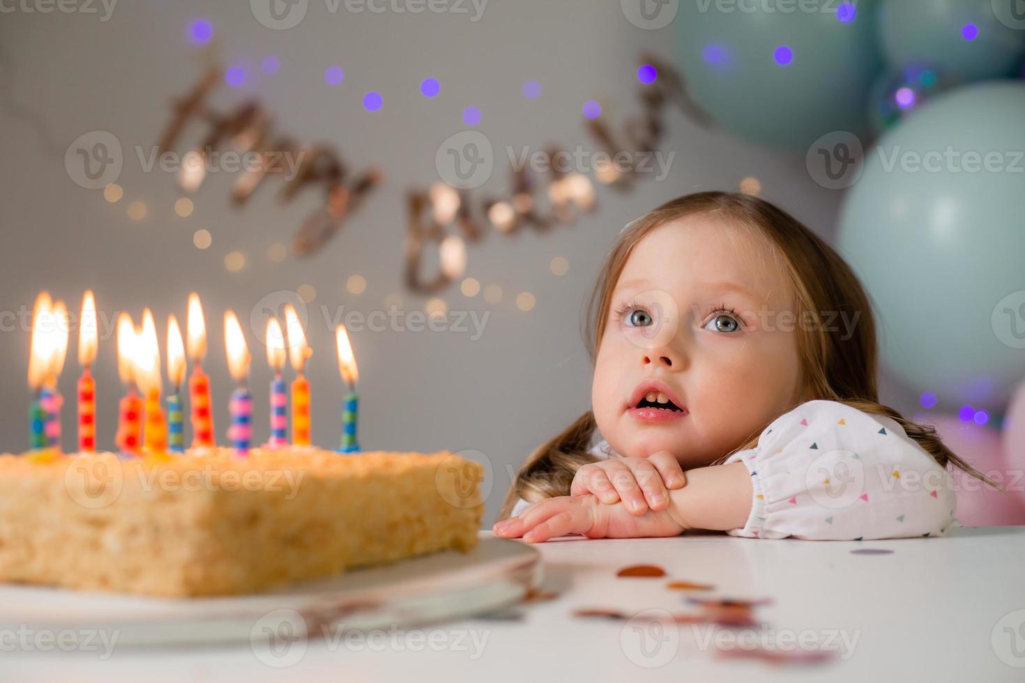 cute little girl blows out candles on a birthday cake at home against a backdrop of balloons. Child's birthday photo