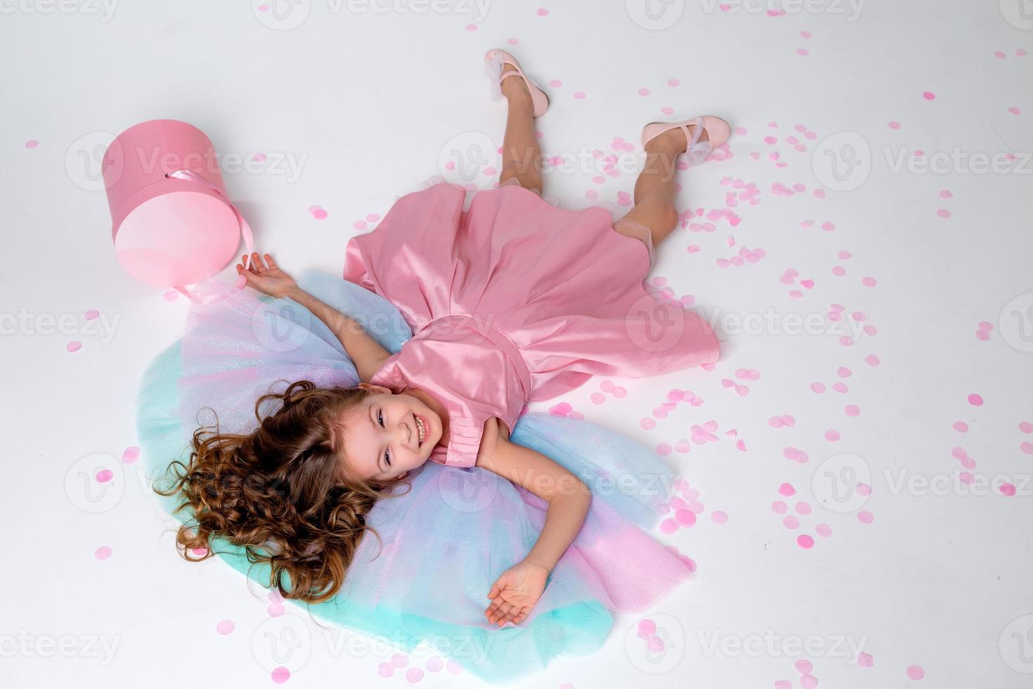 beautiful little girl in a chic pink dress lies on the floor strewn with confetti. top view. fashion and style. child celebrates his birthday. photo in the studio. space for text. High quality photo