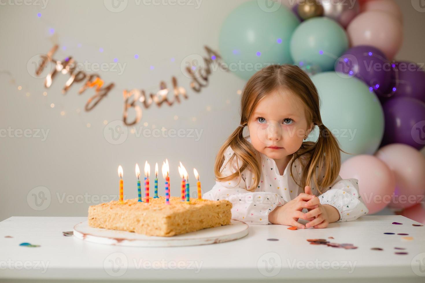 cute little girl blows out candles on a birthday cake at home against a backdrop of balloons. Child's birthday photo