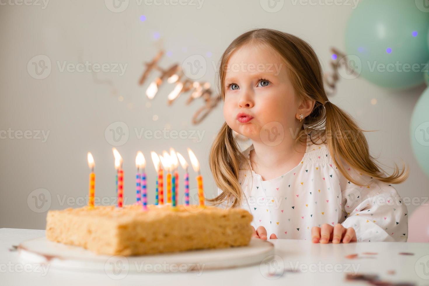 cute little girl blows out candles on a birthday cake at home against a backdrop of balloons. Child's birthday photo