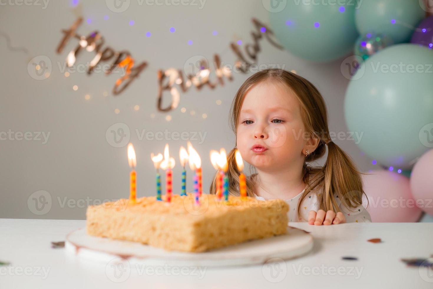 cute little girl blows out candles on a birthday cake at home against a backdrop of balloons. Child's birthday photo