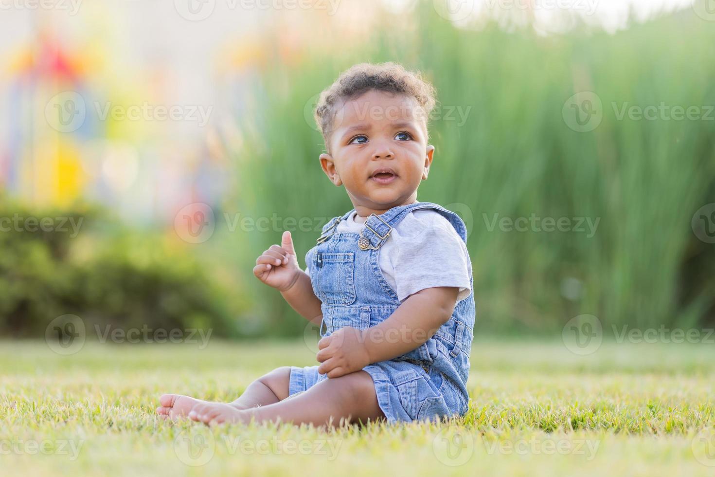 a cute dark-skinned baby in a denim jumpsuit is sitting on the lawn. happy childhood. space for text. High quality photo