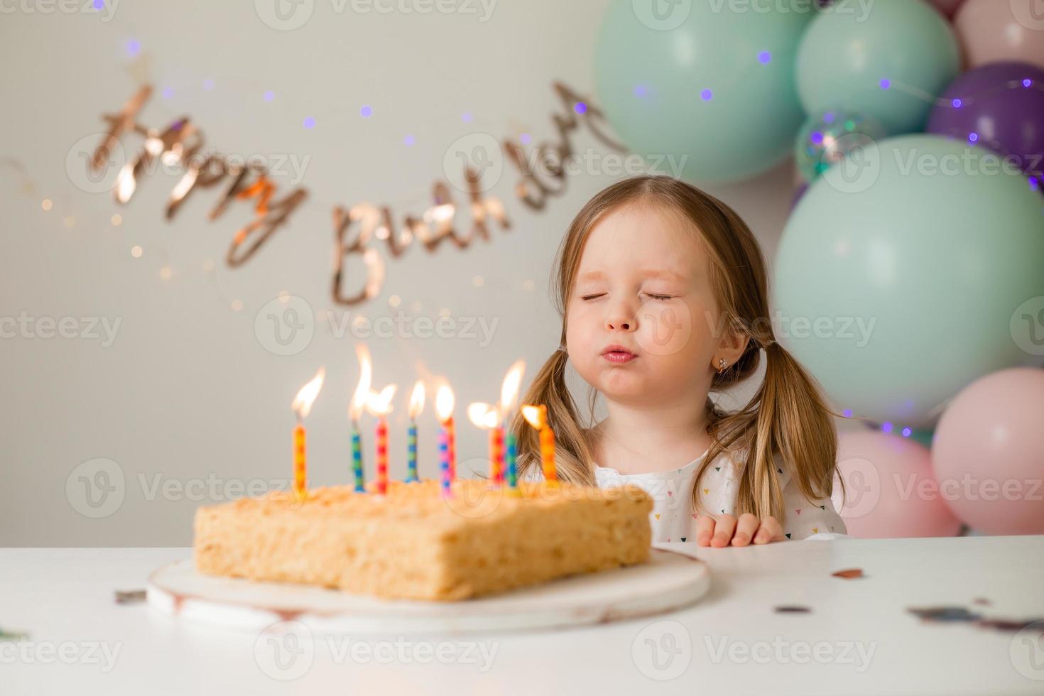 cute little girl blows out candles on a birthday cake at home against a backdrop of balloons. Child's birthday photo