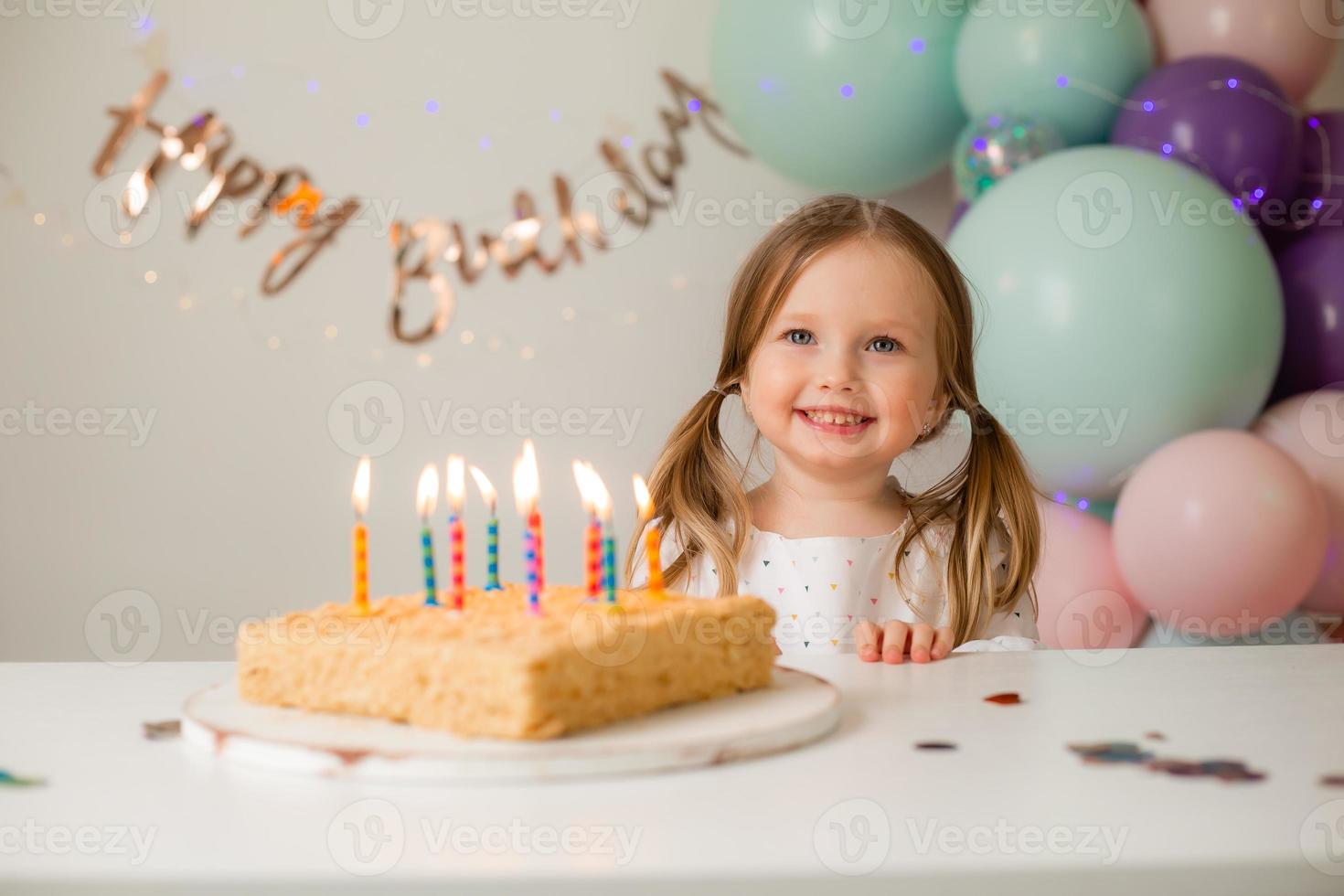 cute little girl blows out candles on a birthday cake at home against a backdrop of balloons. Child's birthday photo