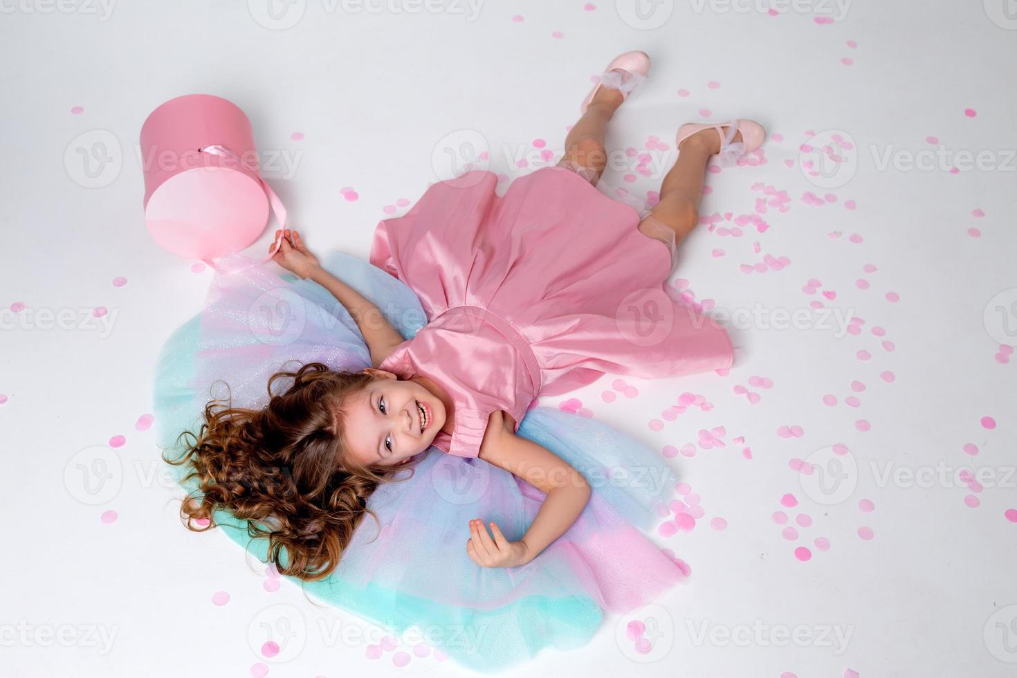 beautiful little girl in a chic pink dress lies on the floor strewn with confetti. top view. fashion and style. child celebrates his birthday. photo in the studio. space for text. High quality photo