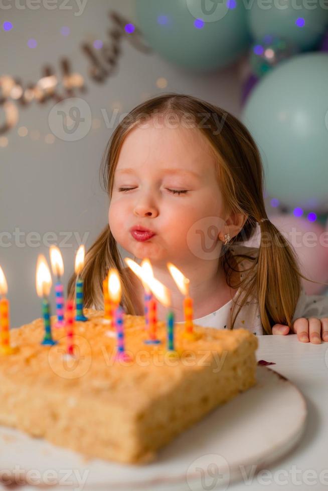 cute little girl blows out candles on a birthday cake at home against a backdrop of balloons. Child's birthday photo