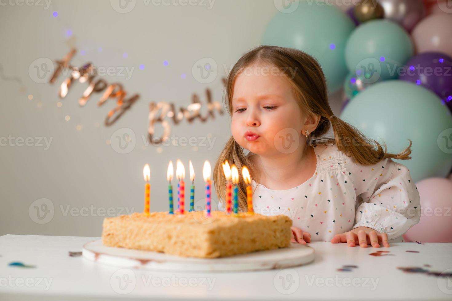 cute little girl blows out candles on a birthday cake at home against a backdrop of balloons. Child's birthday photo