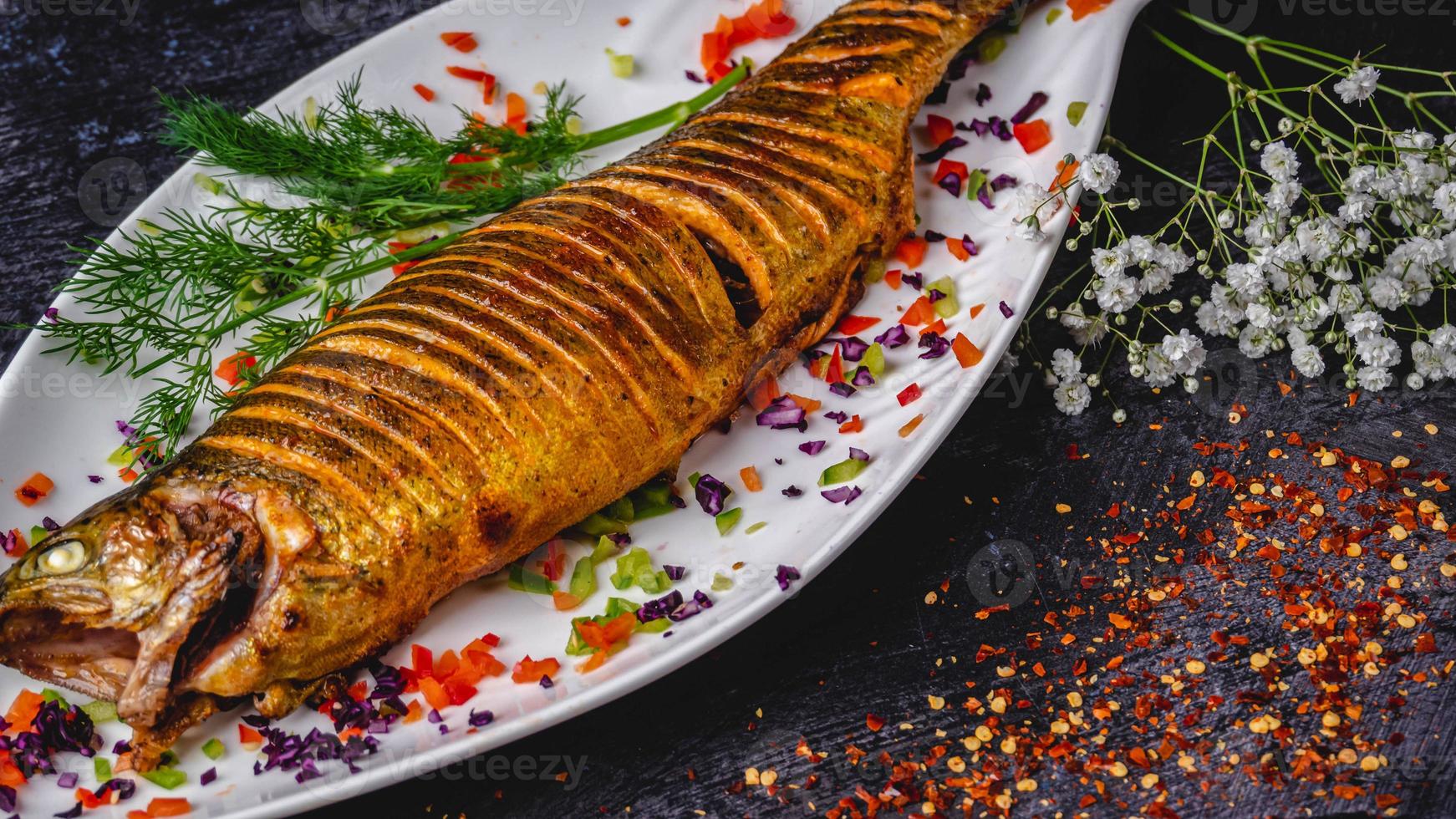 Mouth-watering whole stuffed fish served on a white plate with ginger, tomato, coriander, gypsophila paniculata on the side, and crushed red pepper flakes. photo