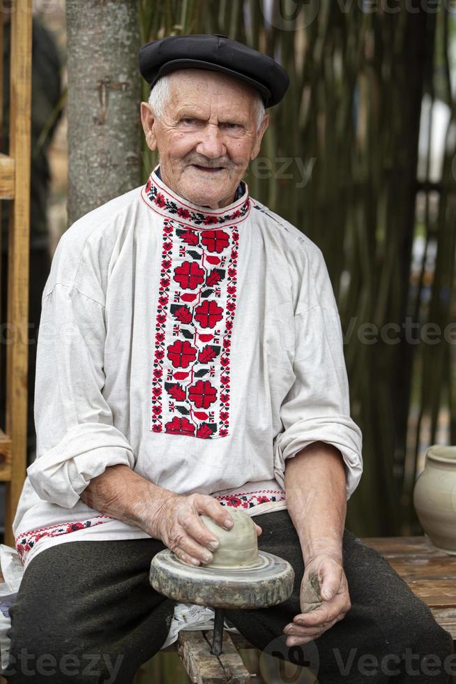 Belarus, the village of Lyaskovichi. August 20, 2022. A holiday of ethnic cultures. An old Slavic man in a Belarusian linen embroidered shirt sculpts items from clay. Ethnic national crafts. photo