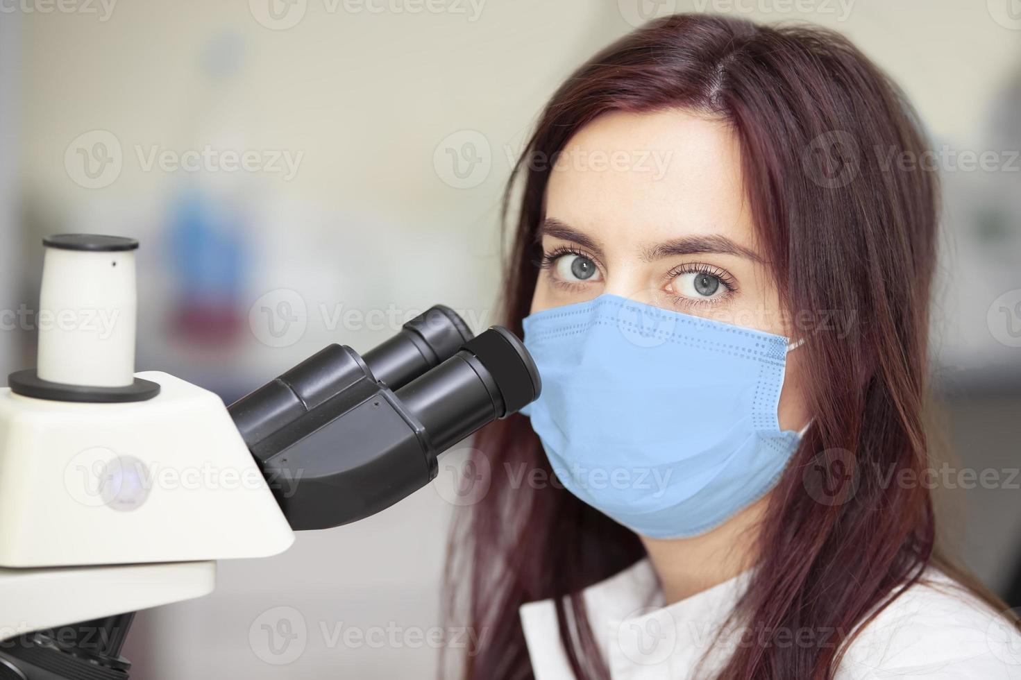 biólogo en un médico máscara con un microscopio. mujer biólogo. laboratorio asistente con un microscopio. foto