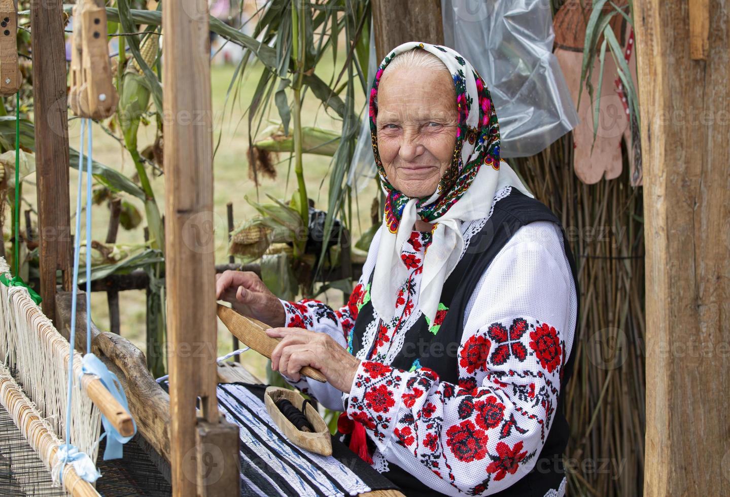 An old Belarusian or Ukrainian woman in an embroidered shirt at a vintage loom. Slavic elderly woman in national ethnic clothes. photo