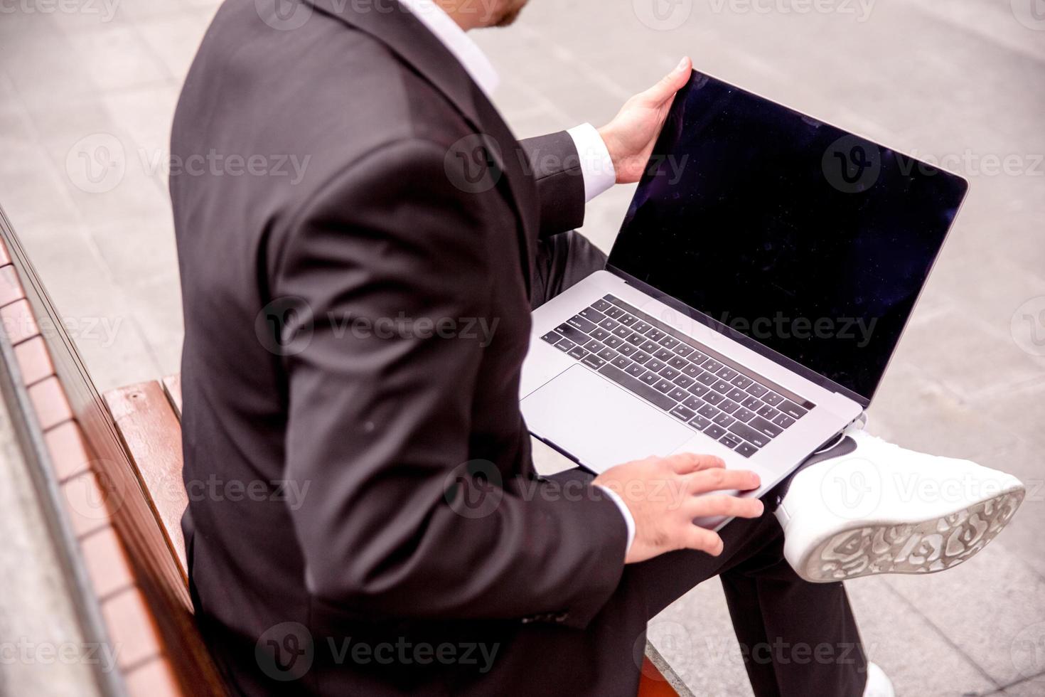 joven chico en un traje trabajos en un computadora en el parque, ordenador portátil cerca arriba foto