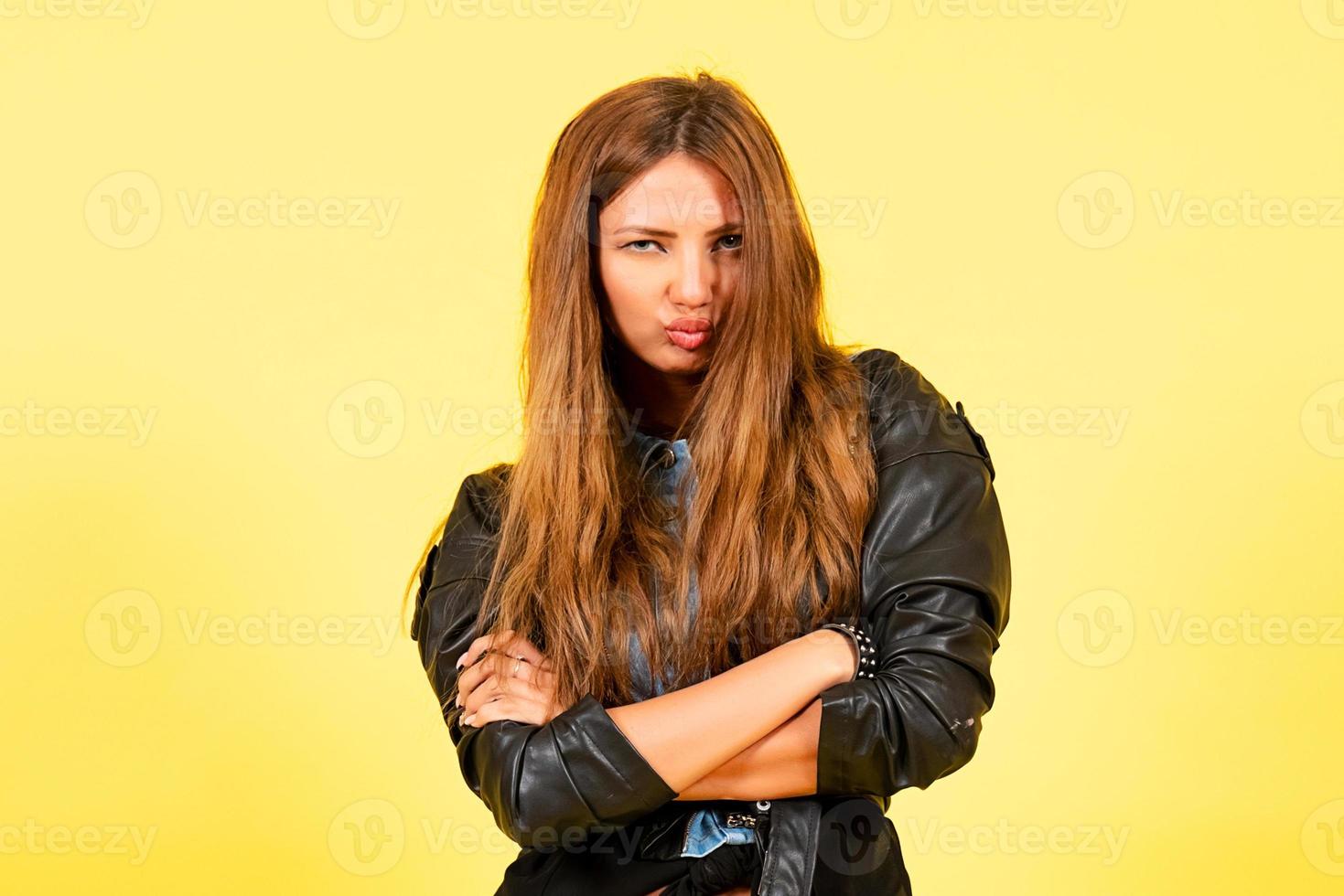 Yellow background, girl in a leather jacket in rock style, shows emotions, free space for text, arms crossed photo