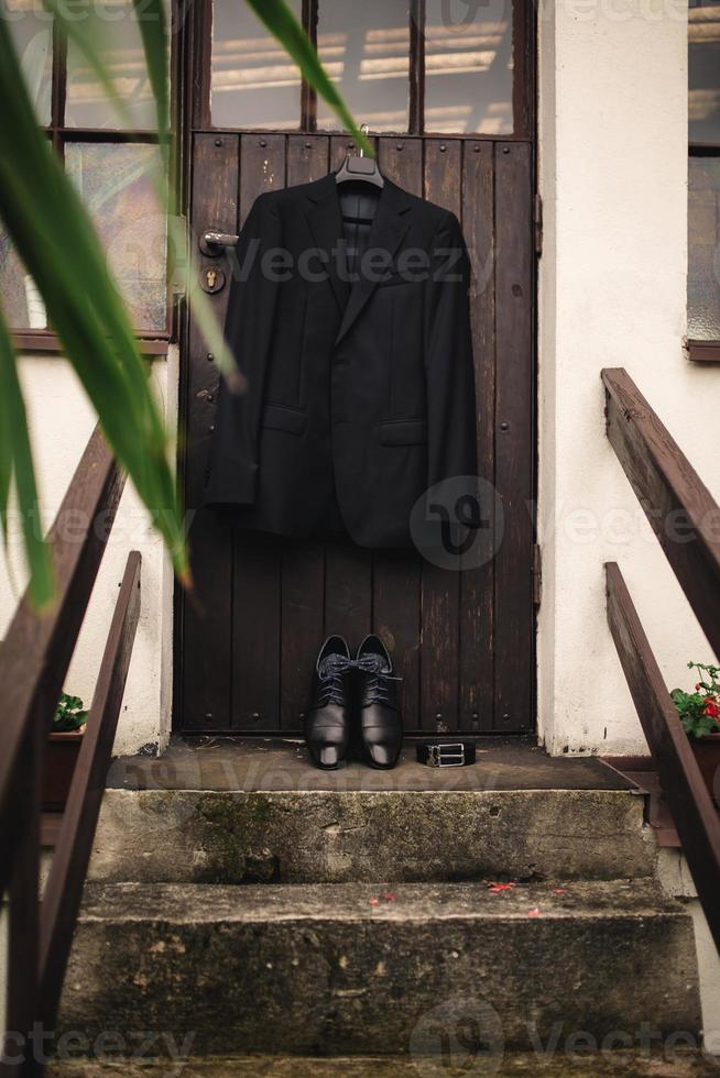 Stylish elegant wedding groom suit with buttonhole hanging on wooden background, copy space. Gray suit hangs above leather groom shoes and pink bowtie. Groom wedding accessories. photo