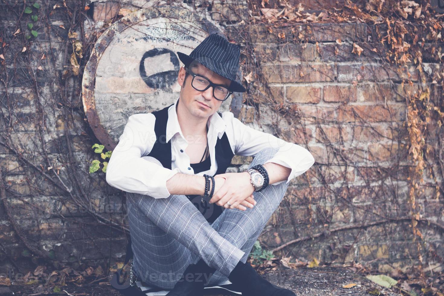 Stylish man with fedora hat relaxing by brick wall outdoors. photo