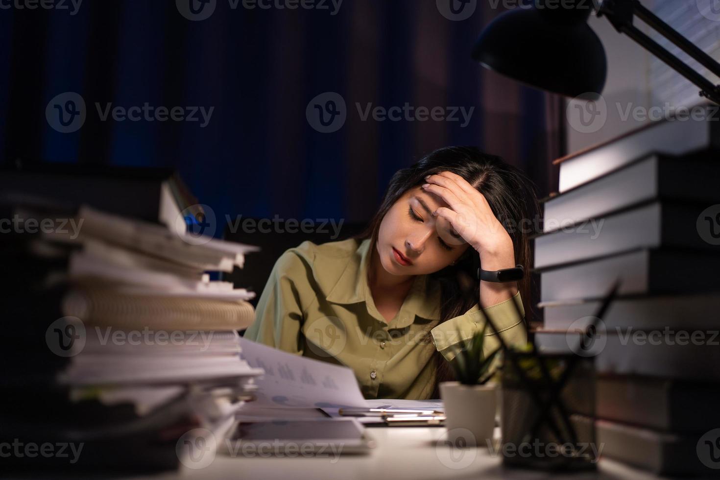 retrato de asiático mujer de negocios sentado y trabajando difícil a con frente de computadora y un montón de documentos en el mesa en lugar de trabajo a tarde con grave acción, trabajo difícil y también tarde concepto foto