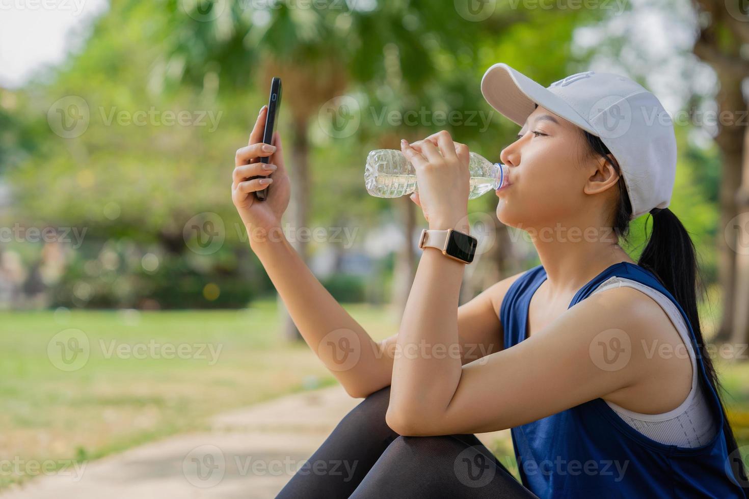 Young beautiful asian drinking water from bottle and using smartphone, she resting after jogging at green park photo