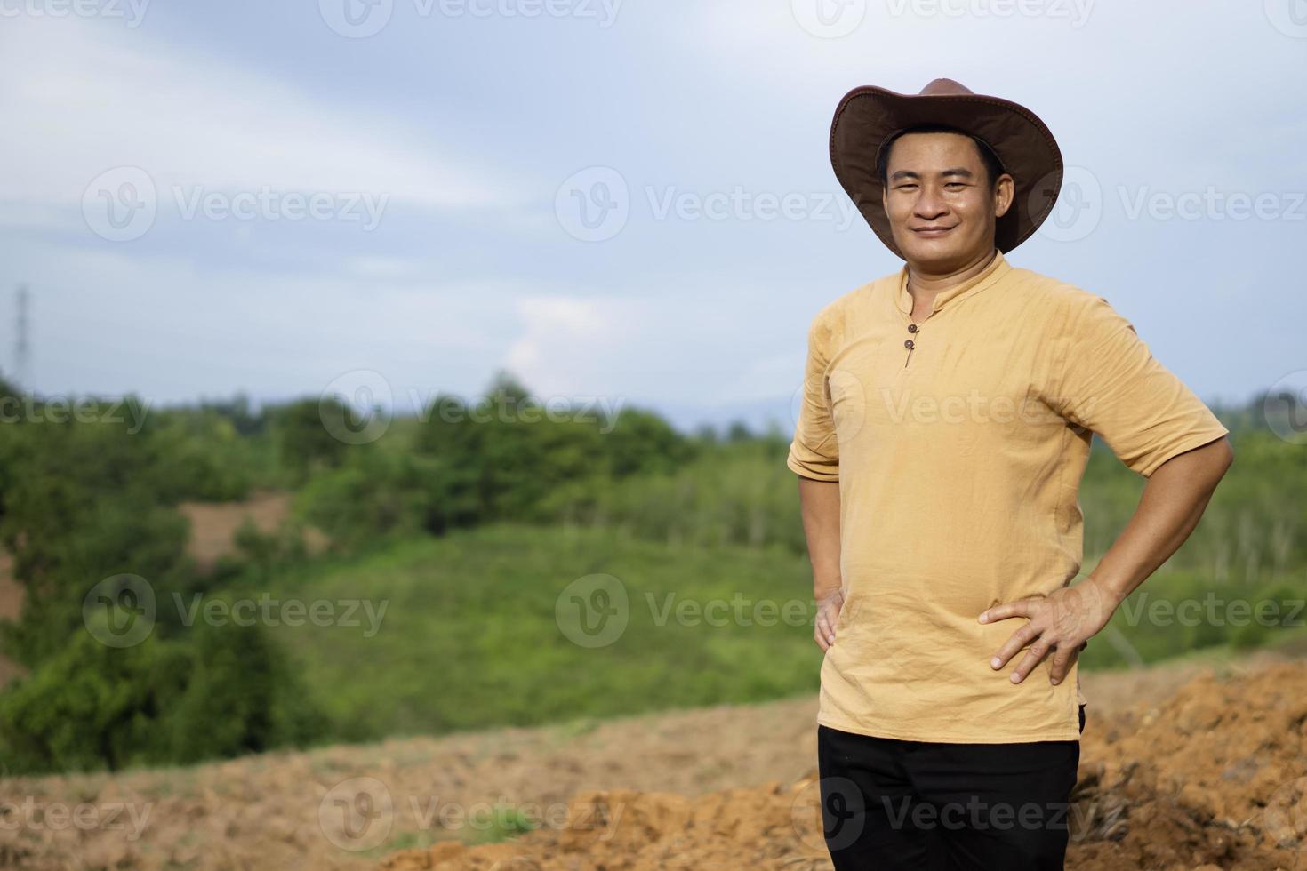 Portrait of Asian man farmer wears hat, brown shirt, puts hands on hips, feels confident, stands at agriculture land  before growing crops. Concept, agriculture occupation. Thai farmer. Copy space. photo