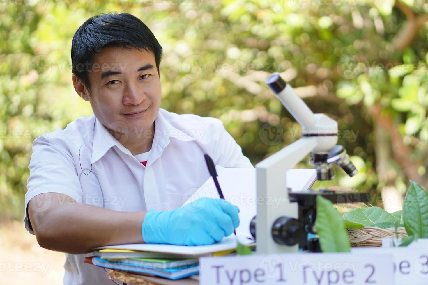 Handsome Asian man science teacher do experiment outdoor, wears blue gloves, take note, use microscope.  Concept, science subject, project work. Experiment, education. Outdoor class. photo