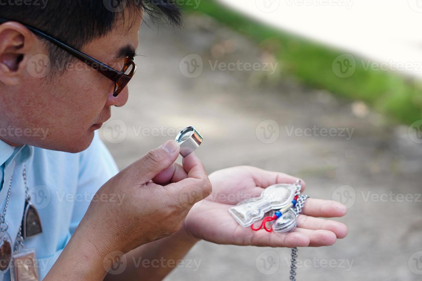 Closeup Asian man holds Buddha amulet and magnifying glass to check details and figure of object. Concept, faith, belief in holy and luck for Buddhists.  Colleting amulet as hobbies, exchange, trade. photo