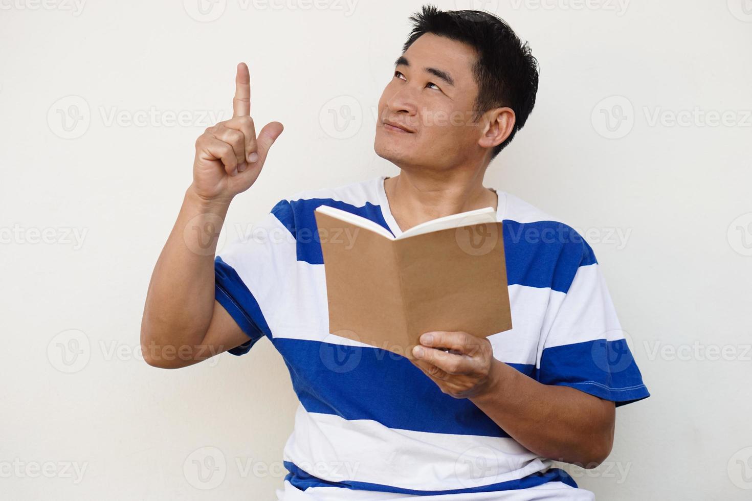 Handsome Asian man holds book and point index finger up. Concept, reading book, get knowledge and idea. Preparing for test. Education. Hobby and pastime. Read for pleasure. Copy space for add text. photo
