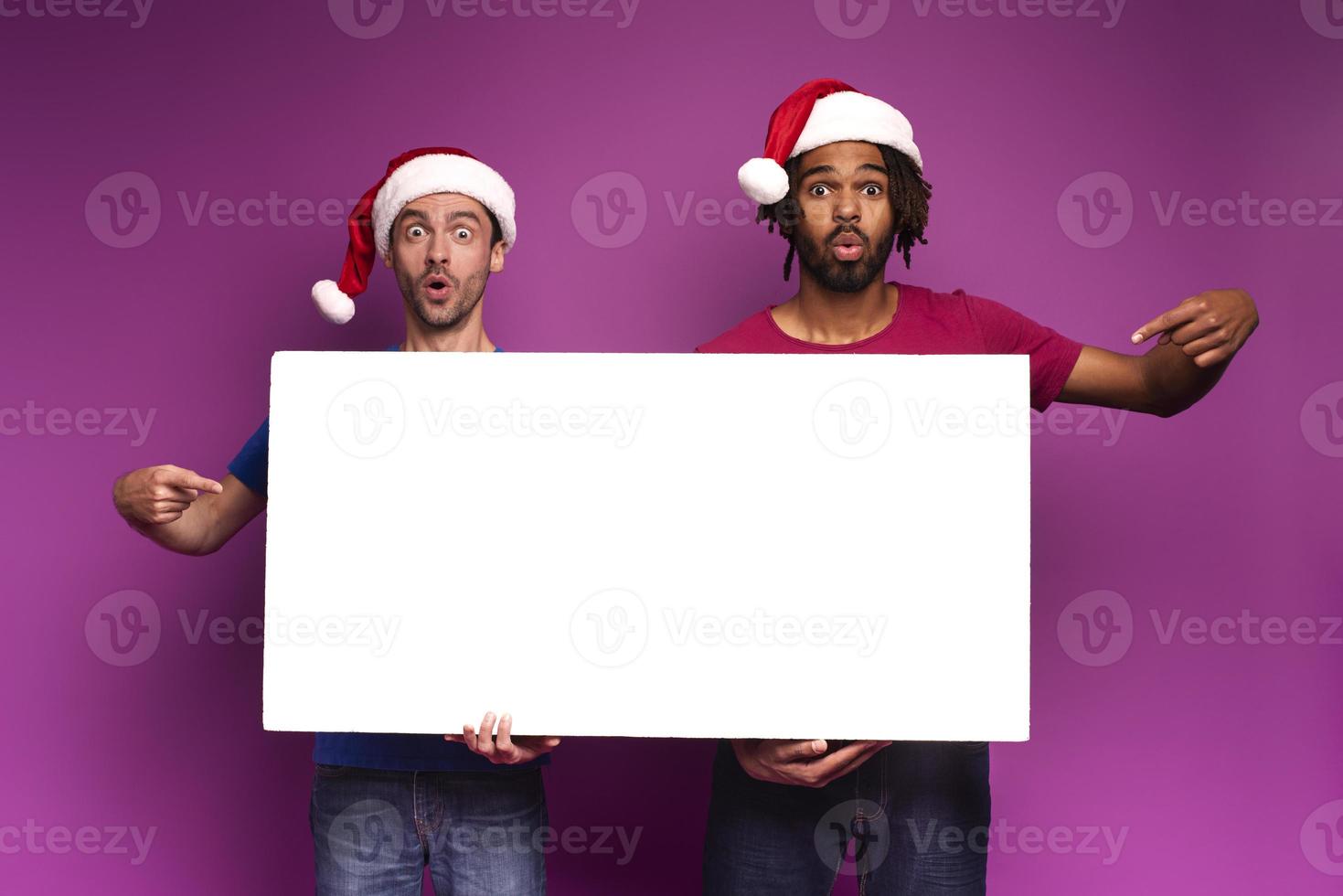 Surprised Amazed friends with Christmas hat hold a white banner for your message. Purple background photo