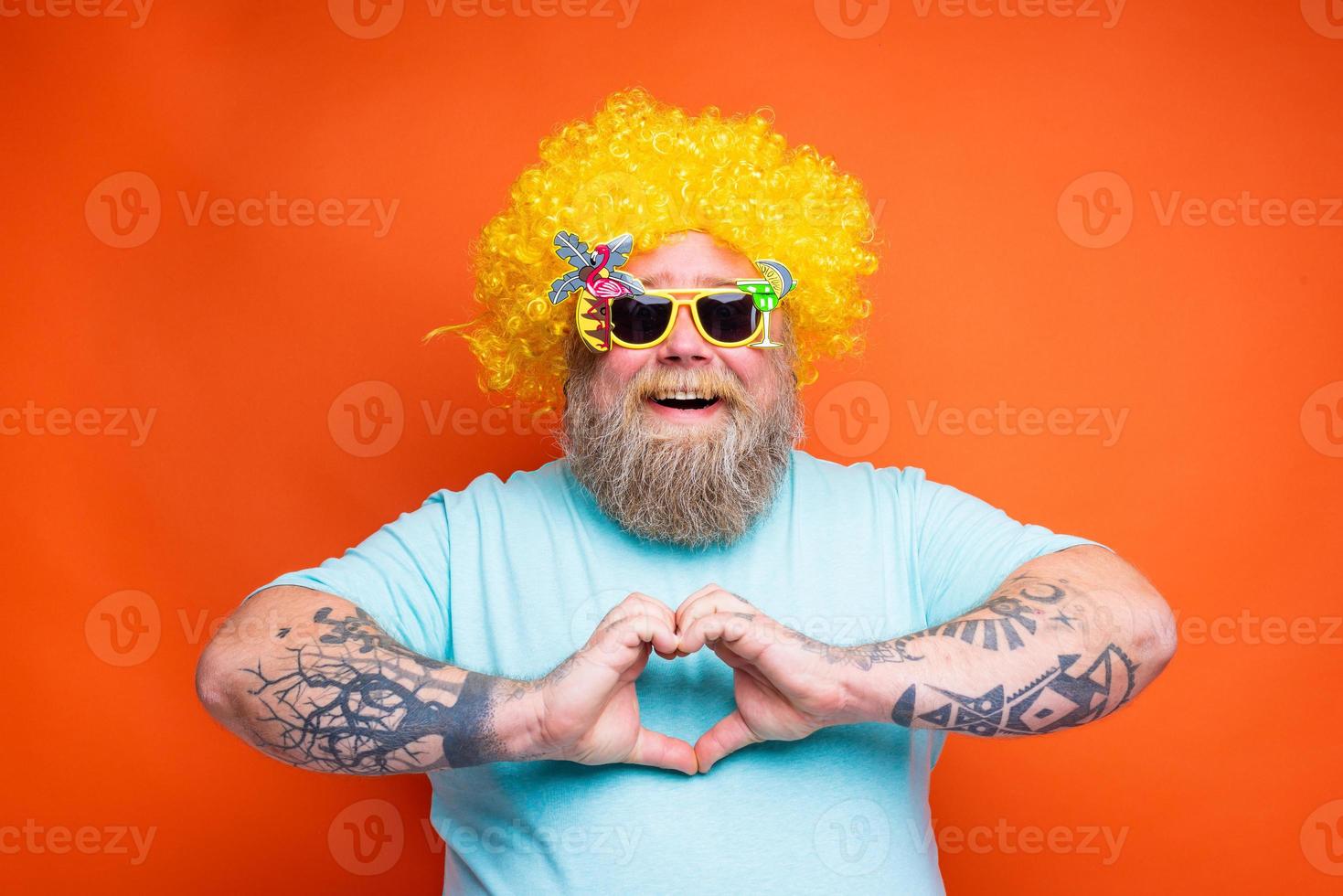 grasa contento hombre con barba, tatuajes y Gafas de sol hace corazón forma con manos foto