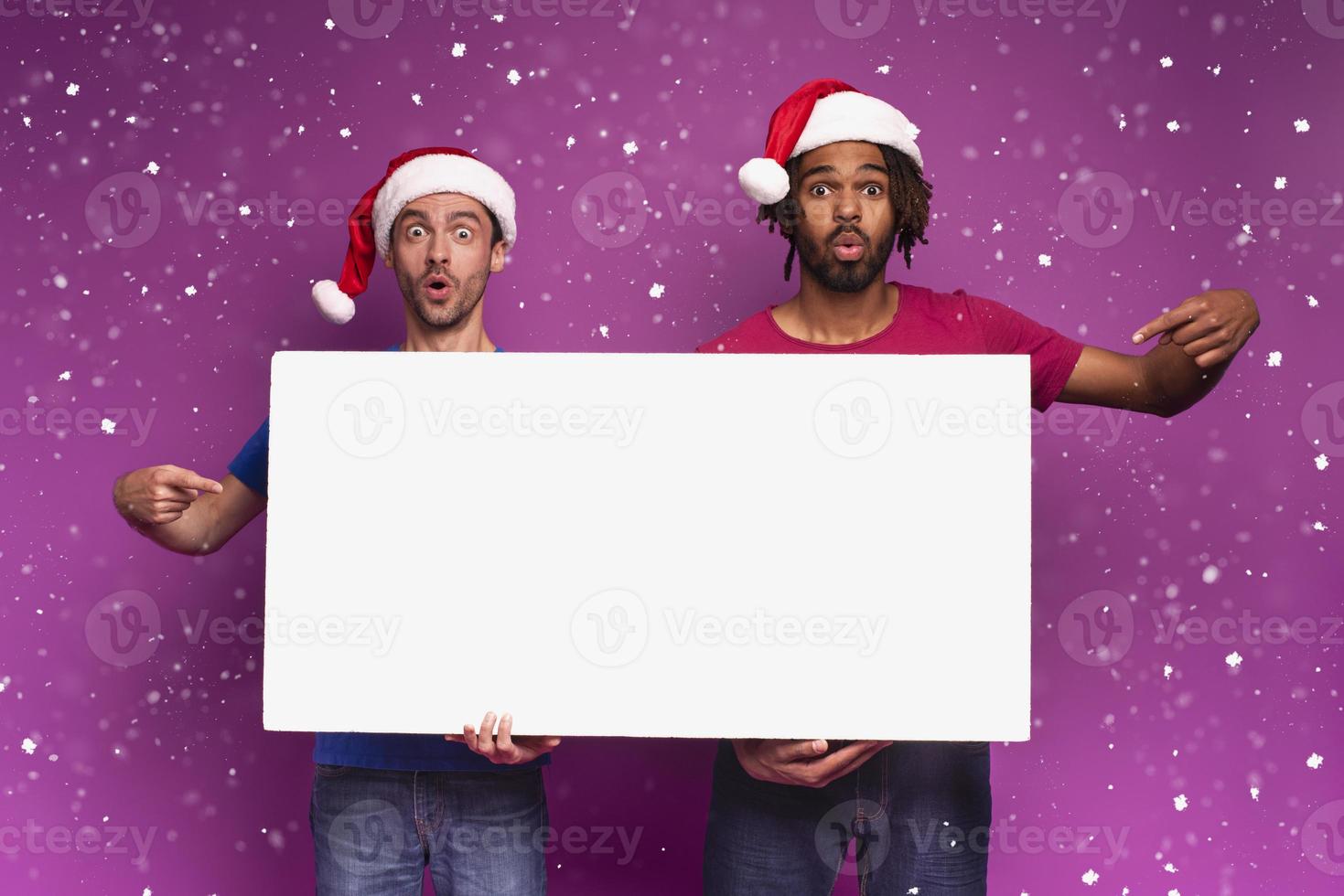 Surprised Amazed friends with Christmas hat hold a white banner for your message. Purple background photo