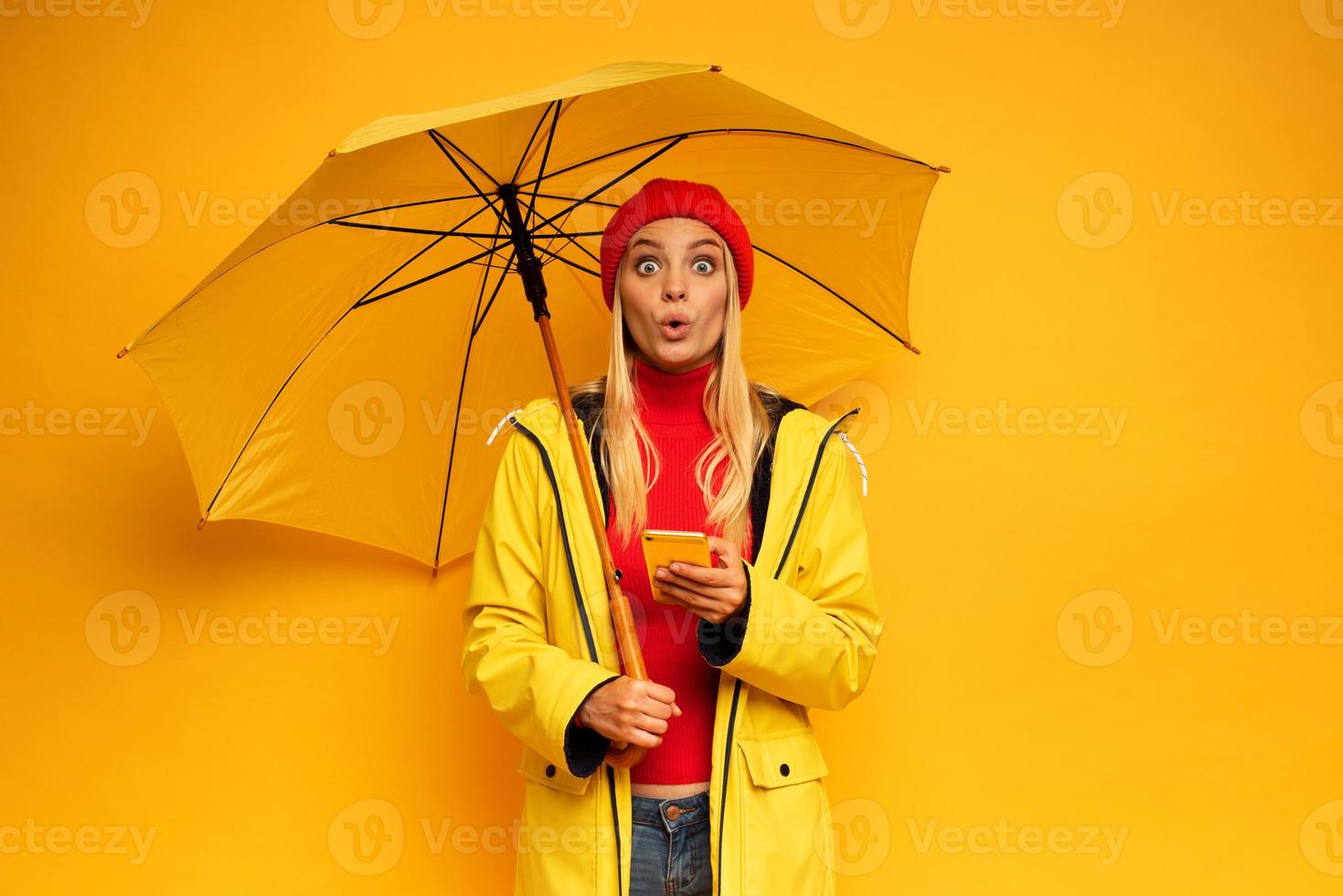 Girl with smartphone and umbrella on yellow background surprised for the weather photo