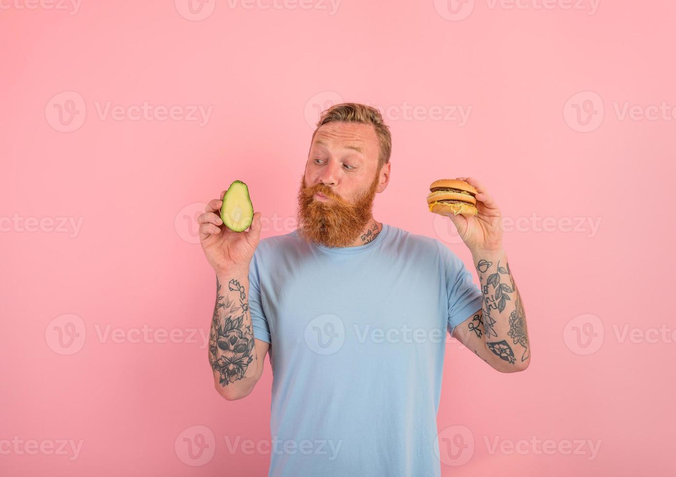 Thoughtful man with beard and tattoos is undecided if to eat an avocado or an hamburger photo