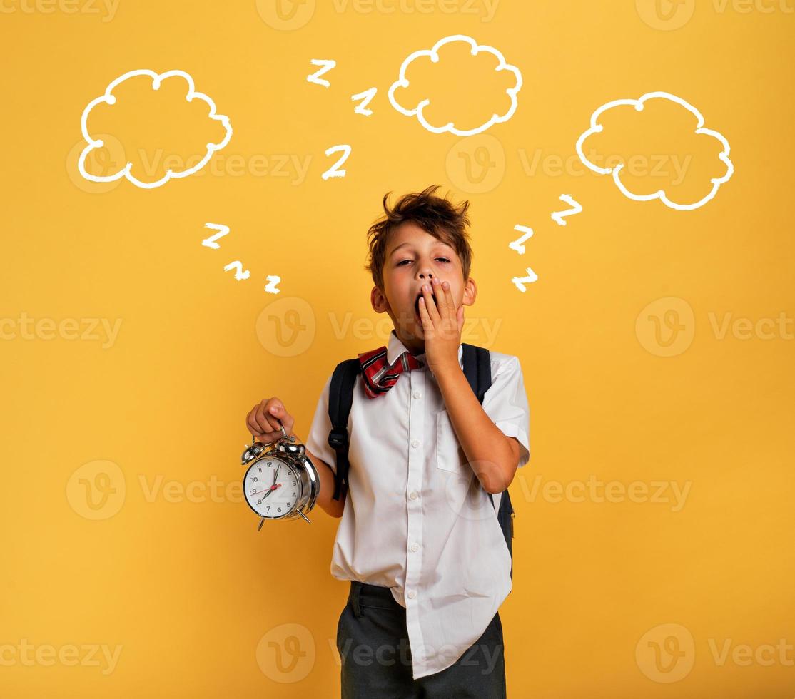 Young child student with ringing alarm clock is lazy going to school. Yellow background. photo