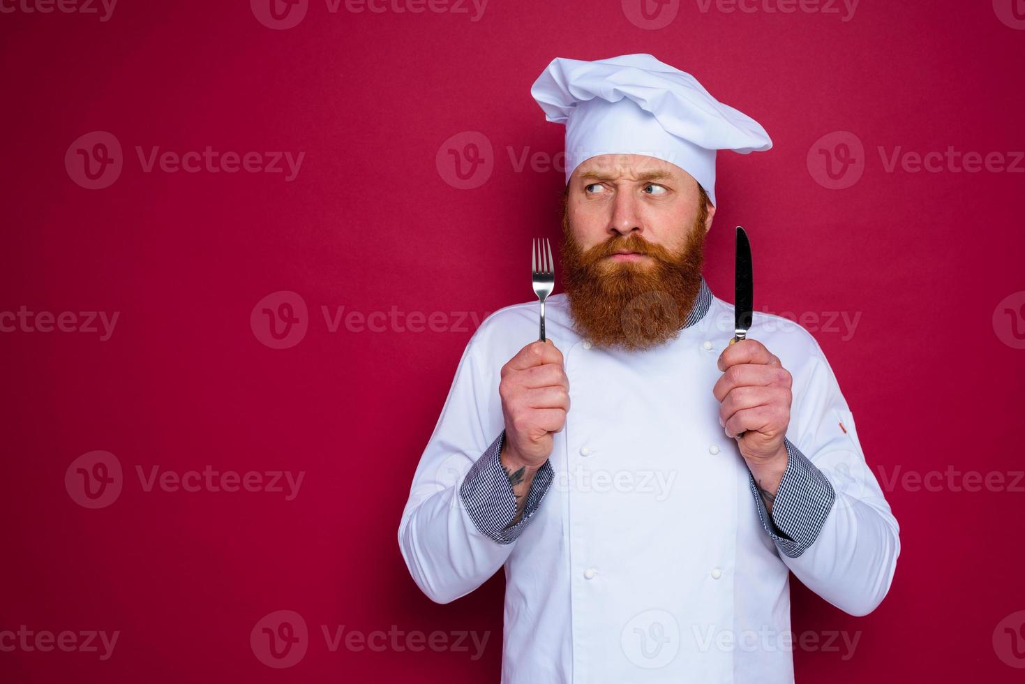 pensive chef with beard and red apron holds cutlery in hand photo